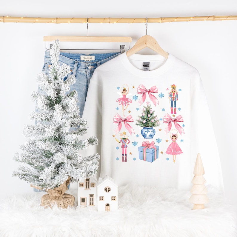 Op de afbeelding: Witte kersttrui met een feestelijk ontwerp met een ballerina, een notenkraker, een kerstboom en een cadeaudoos. Het ontwerp is in roze, blauw en goud.