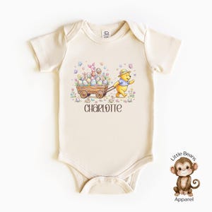May include: Cream-colored baby onesie with an Easter-themed graphic. The illustration features Winnie the Pooh pulling a wooden cart filled with Easter eggs, alongside Piglet. The name "CHARLOTTE" is printed below the graphic. The onesie has short sleeves and snap closures.