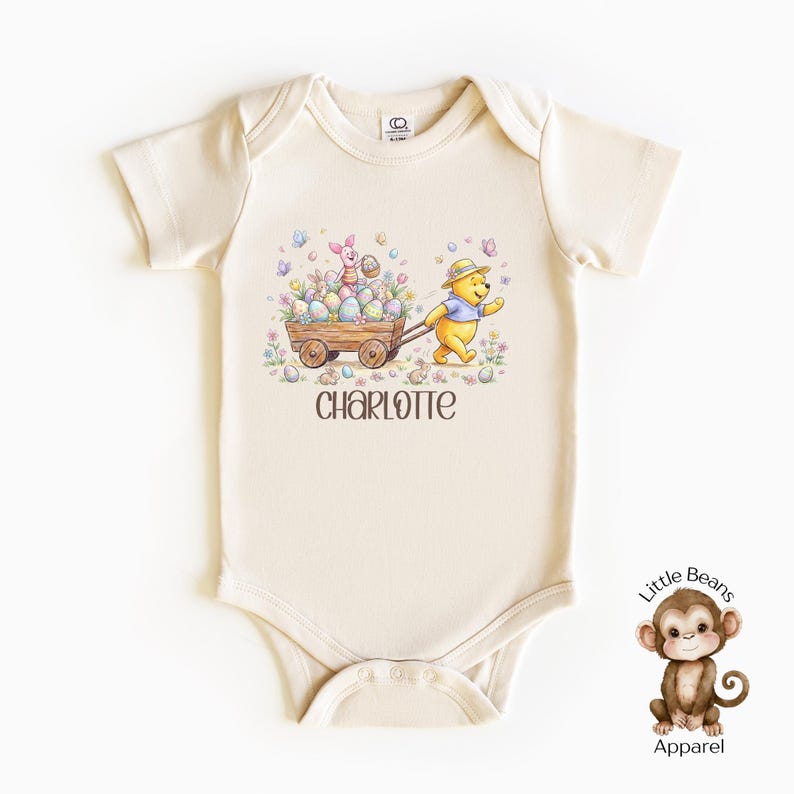 May include: Cream-colored baby onesie with an Easter-themed graphic. The illustration features Winnie the Pooh pulling a wooden cart filled with Easter eggs, alongside Piglet. The name "CHARLOTTE" is printed below the graphic. The onesie has short sleeves and snap closures.
