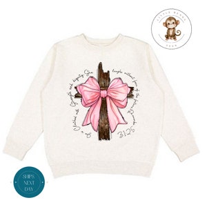 May include: A white long-sleeved sweatshirt with a pink bow and a wooden cross. The text "She is Clothed with Strength and Dignity. She laughs without fear of the future. 3125" is printed around the cross.