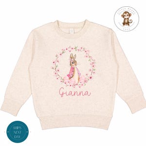 May include: A beige sweatshirt featuring a pink floral wreath with a bunny rabbit in the centre. The name "Gianna" is printed below the wreath.