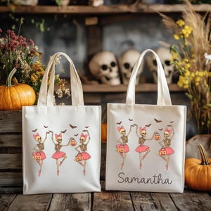 Puede incluir: Dos bolsas de mano de color crema con asas color crema. Cada bolsa presenta un diseño de Halloween de bailarinas esqueléticas bailando con tutús rosas, sosteniendo calabazas. Una bolsa tiene el nombre "Samantha".