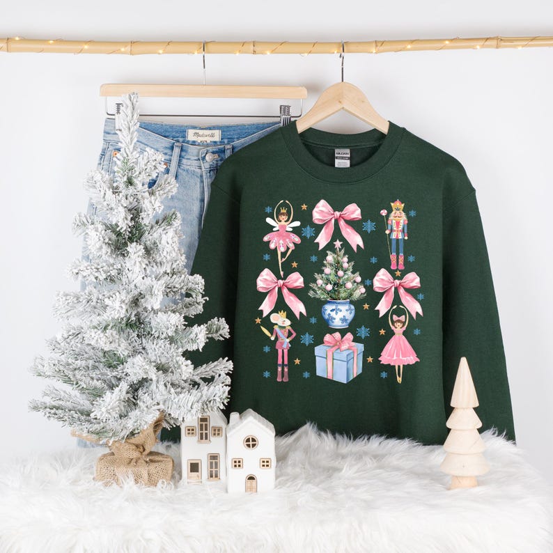 Op de afbeelding: Een donkergroene kersttrui met een feestelijk ontwerp met een notenkraker, een ballerina, een kerstboom en roze strikken.