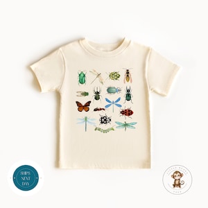 May include: A cream coloured t-shirt with a colourful bug and insect print. The shirt has short sleeves and a crew neck.