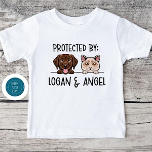 May include: White t-shirt with a brown dog and a tan cat peeking over a white bar. The text "PROTECTED BY: LOGAN & ANGEL" is printed on the shirt.