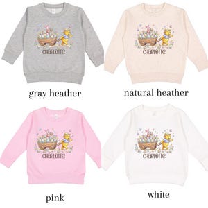 May include: Four children's sweatshirts in gray heather, natural heather, pink, and white. Each features a graphic of a bear pushing a cart filled with Easter eggs, with the name "Charlotte" below. The sweatshirts are long-sleeved.