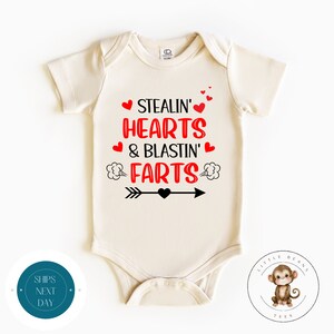 May include: A cream-colored baby onesie with a red and black graphic that says "Stealin' Hearts & Blastin' Farts" with a black arrow and a red heart in the middle.