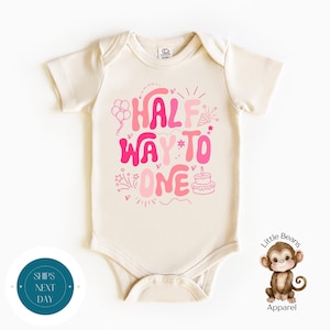 May include: Cream-colored baby onesie with the text "HALF WAY TO ONE" in shades of pink. The design includes balloons, a party hat, stars, and a cake. The onesie is a short-sleeved garment.