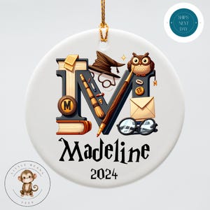 May include: A white ceramic ornament with a black letter M decorated with a witch's hat, glasses, a quill, and an owl. The name Madeline and the year 2024 are written below the letter.