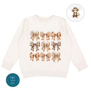 May include: A cream-colored sweatshirt with a pattern of brown and beige bows. The sweatshirt has long sleeves and a crew neck.