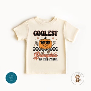 Op de afbeelding: Een licht beige t-shirt met een vintage-stijl afbeelding van een pompoen met een zonnebril en de tekst "Coolest Pumpkin in the Patch".