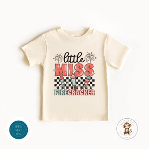 Puede incluir: Una camiseta de color crema con un diseño gráfico de fuegos artificiales y el texto "little miss firecracker".
