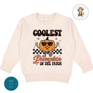 Könnte beinhalten: Ein beiger Pullover mit einem Aufdruck einer lächelnden Kürbis, der eine Sonnenbrille trägt und den Text "Coolest Pumpkin in the Patch".