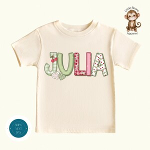 May include: A cream-coloured t-shirt featuring the name "JULIA" in decorative letters. The letters are adorned with cherries, hearts, and floral patterns. The shirt has a round neckline and short sleeves. A small monkey logo is in the top right corner.