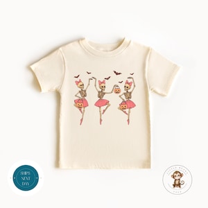 May include: Cream-colored t-shirt featuring a Halloween-themed graphic. The design shows three skeleton ballerinas in pink tutus, holding pumpkin buckets, and wearing pink bows. Bats are flying above them.