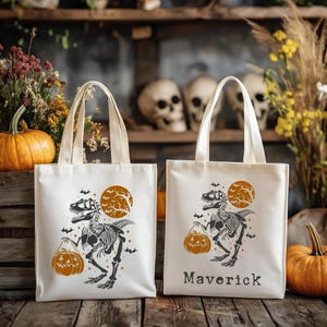 May include: Two off-white tote bags with cream-colored handles. Each bag features a black dinosaur skeleton holding an orange pumpkin, with a full orange moon in the background. One bag has the name "Maverick" printed in black.