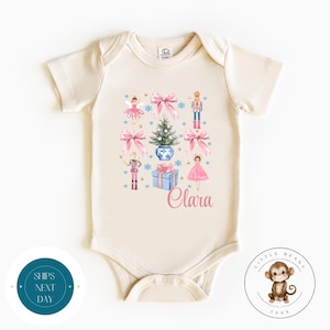 Puede incluir: Un body de bebé de color crema con un diseño navideño que incluye una bailarina, un cascanueces, un árbol de Navidad y una caja de regalo. El body tiene el nombre "Clara" impreso en él.