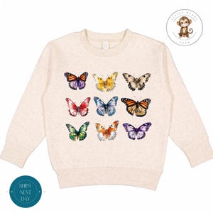 May include: A beige long-sleeved sweatshirt with a colorful butterfly pattern. The butterflies are arranged in a grid pattern and are all different colors and sizes.