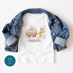May include: White t-shirt with a colorful graphic featuring Winnie the Pooh pulling a wooden cart filled with Easter eggs, with Piglet in the cart. The name "Charlotte" is printed below the graphic. A blue denim jacket is draped over the shirt.