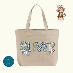 May include: Beige canvas tote bag with the name "OLIVER" in colorful baseball-themed letters. The letters feature baseballs, bats, and stars. A small cartoon monkey with the text "Little Beans Apparel" is in the upper right corner.