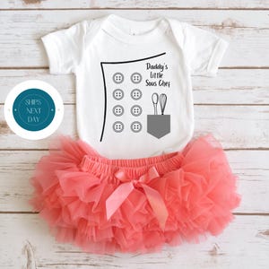 May include: White baby onesie with a black chef's hat design and the text "Daddy's Little Sous Chef". The hat has buttons and a pocket with a spoon and whisk. The onesie is paired with a coral tulle tutu.