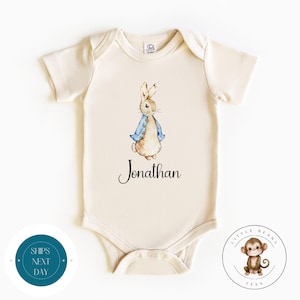 May include: A cream colored baby onesie with a watercolor illustration of Peter Rabbit wearing a blue jacket. The onesie has the name "Jonathan" printed in black cursive lettering.
