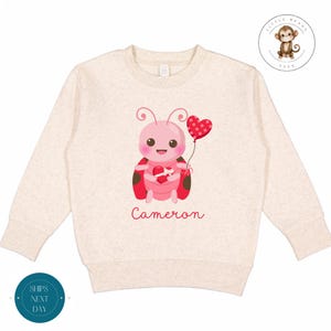 Pode incluir: Uma camisola bege claro com uma joaninha rosa segurando um balão vermelho em forma de coração. A joaninha tem manchas pretas e um laço vermelho. O texto "Cameron" está impresso abaixo da joaninha.