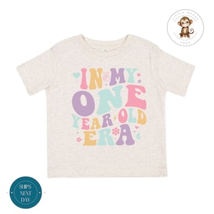 May include: A light beige toddler t-shirt with a colorful graphic that says "In My One Year Old Era" with peace sign, hearts, and flowers.