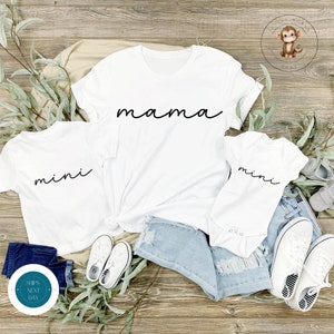 Op de afbeelding: Witte T-shirts met de woorden "mama" en "mini" in zwarte schrift. De shirts liggen op een houten oppervlak met een blauwe jeans en witte sneakers.