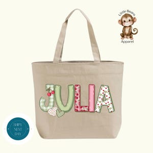 May include: A beige canvas tote bag displaying the name "JULIA" in colourful letters. The letters feature cherry and gingham patterns. A small monkey logo is in the upper right corner. The words "Ships Next Day" are on a blue circle.
