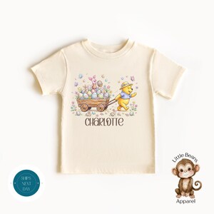 May include: Cream-colored toddler t-shirt featuring an Easter-themed graphic. The design includes Winnie the Pooh pulling a wooden cart filled with Easter eggs, with Piglet in the cart. The name "Charlotte" is printed below the graphic.