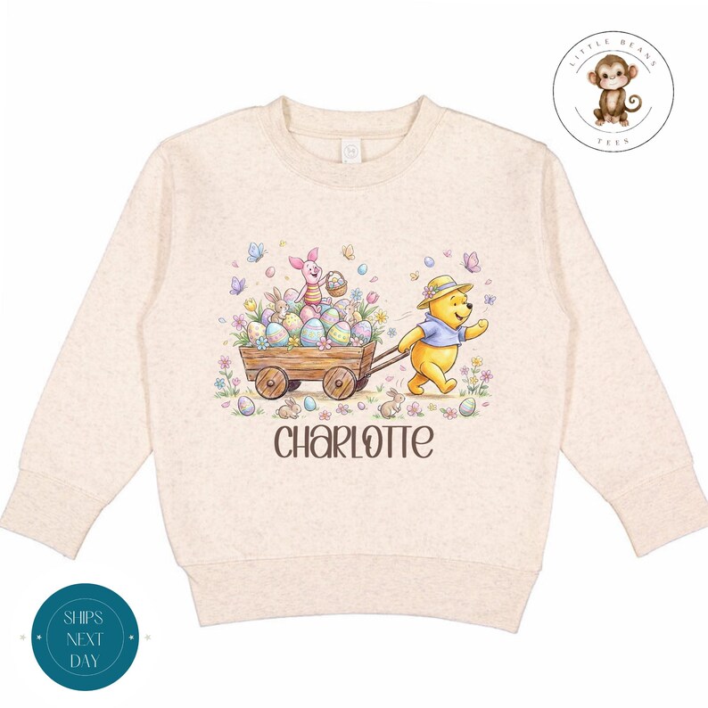 May include: A beige long-sleeved sweatshirt featuring a colorful Easter-themed graphic. The design includes Winnie the Pooh, Piglet, and a wooden cart filled with Easter eggs. The name "CHARLOTTE" is printed below the graphic.