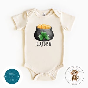 May include: A cream coloured baby onesie with a pot of gold illustration. The pot is filled with gold coins and has a shamrock on top. The text "CAIDEN" is printed below the pot.