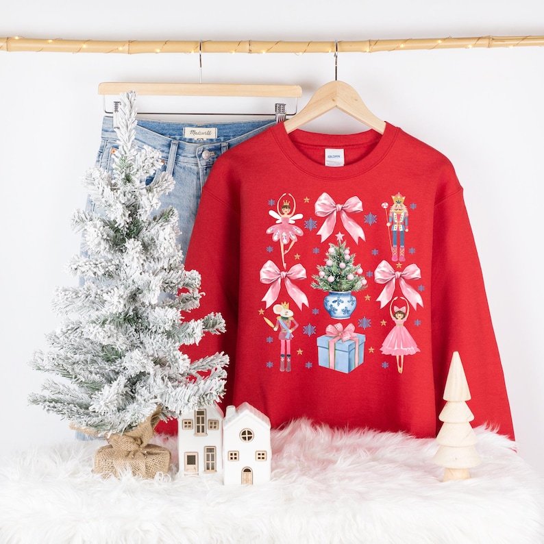 Op de afbeelding: Rode kersttrui met een feestelijk ontwerp met een kerstboom, notenkraker, ballerina en strikken.