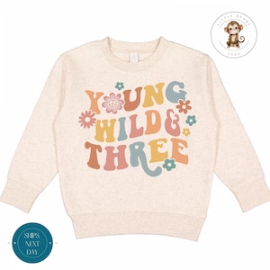 Op de afbeelding: Een beige longsleeve sweatshirt met de tekst "Young Wild & Three" in een retro lettertype met kleurrijke bloemen en een vredesteken.