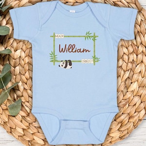 May include: Light blue baby onesie with the name "William" in brown script, framed by bamboo stalks and leaves. A small panda illustration is below the name. The text "MAR 2027" is also included. The onesie is on a woven mat.