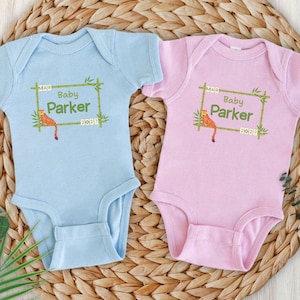 May include: Two baby onesies, one light blue and one pink, are displayed on a woven mat. Each onesie features a green bamboo frame with the text "Baby Parker" and a small tiger illustration. The date "2027" is also printed.