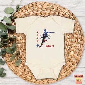 May include: Cream-colored baby onesie featuring a soccer-themed graphic. The design includes the numbers "2026", a soccer player silhouette, and the text "Dallas, TX". The graphic incorporates red, white, and blue colors.