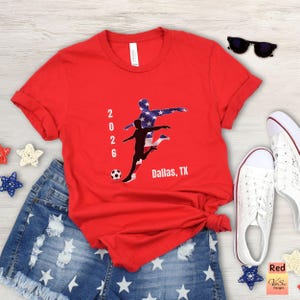 May include: A red t-shirt with a soccer-themed graphic featuring two players, a soccer ball, and the text "2026 Dallas, TX." The design includes a silhouette of a player with the American flag pattern. The shirt is paired with denim shorts and white sneakers.
