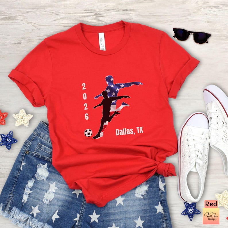 May include: A red t-shirt with a soccer-themed graphic featuring two players, a soccer ball, and the text "2026 Dallas, TX." The design includes a silhouette of a player with the American flag pattern. The shirt is paired with denim shorts and white sneakers.