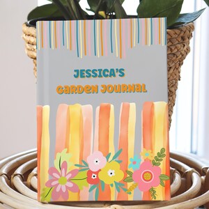Puede incluir: Un diario titulado "Jessica's Garden Journal" con fondo gris. La portada presenta rayas verticales coloridas e ilustraciones florales en rosa, amarillo y naranja. El diario se muestra frente a una planta.