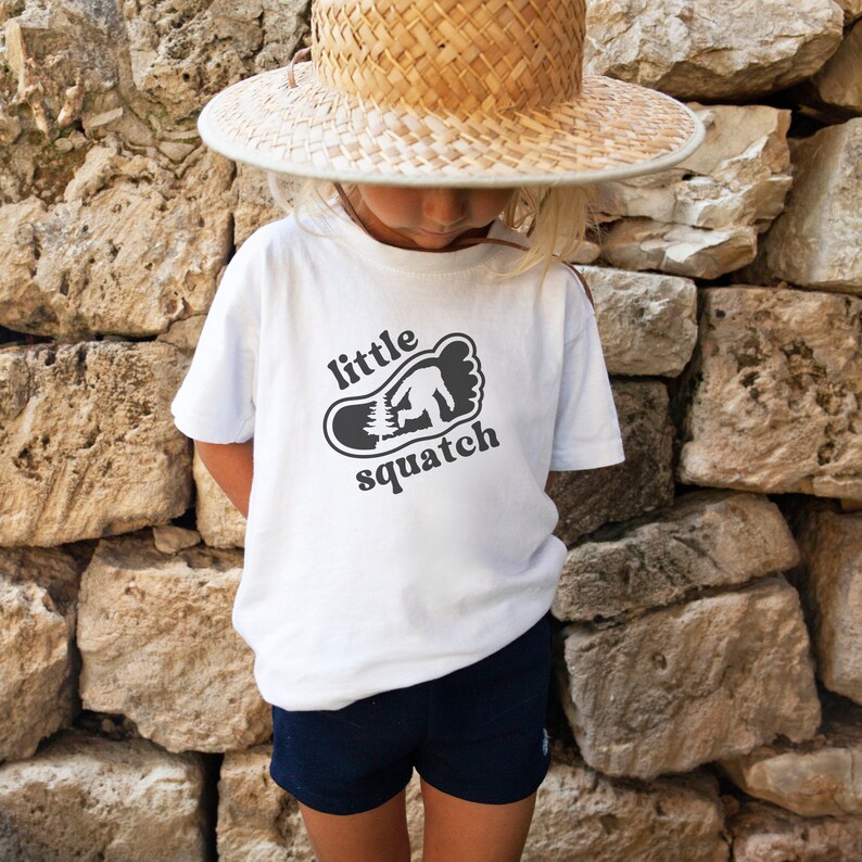 May include: White t-shirt with a black graphic of a footprint with a small figure inside. The text "little squatch" is printed above the footprint.
