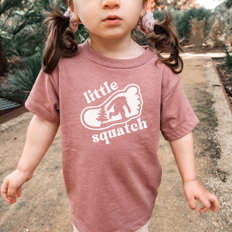 May include: A mauve colored t-shirt with a white graphic of a footprint with a small tree and a bigfoot inside. The text "little squatch" is printed below the footprint.
