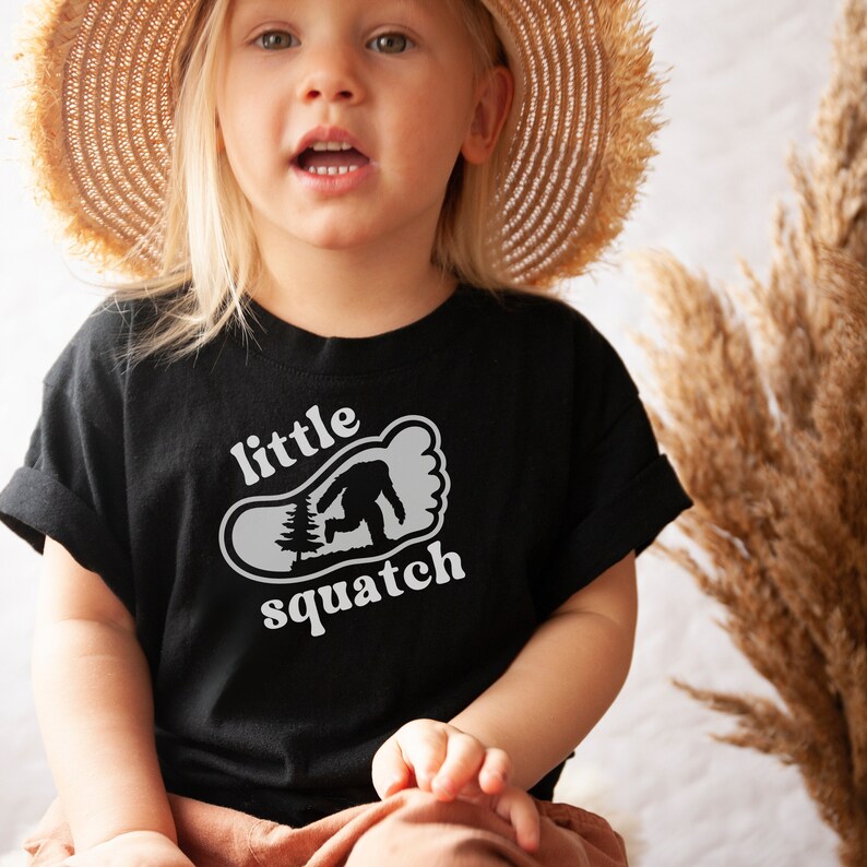 May include: A child wearing a black t-shirt with a white graphic of a footprint with a small tree and a Bigfoot inside. The text "little squatch" is printed below the footprint. The child is wearing a straw hat and is sitting down.