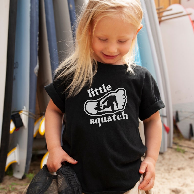May include: A black t-shirt with a white graphic of a footprint with a small tree and a Bigfoot inside. The text "little squatch" is printed below the footprint.