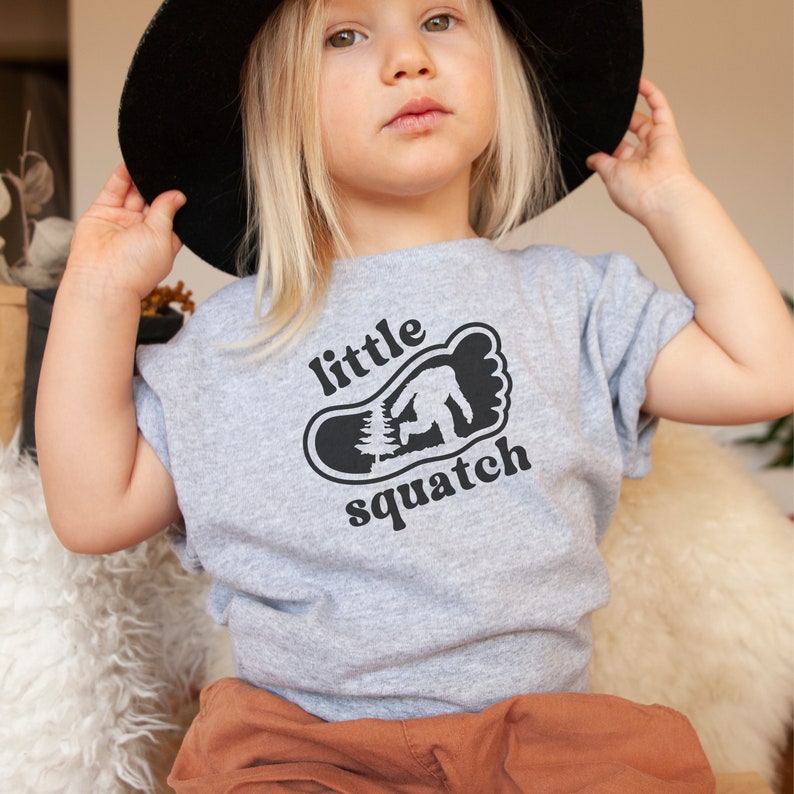 May include: A gray t-shirt with a black graphic of a footprint with a small tree and a Bigfoot inside. The text "little squatch" is printed below the footprint.