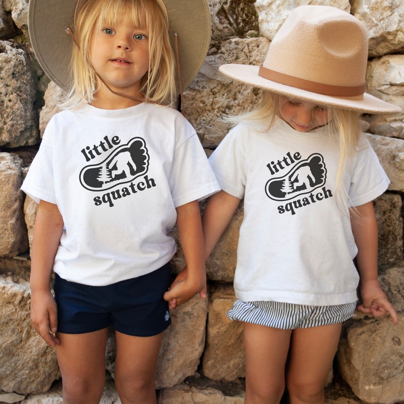 May include: Two children wearing white t-shirts with a black footprint graphic and the text "little squatch". The children are wearing hats and shorts.