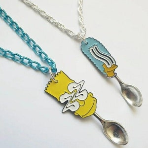 Może przedstawiać: Dwa nowatorskie naszyjniki z wisiorkami z postaciami z kreskówek przymocowanymi do łyżek. Jeden wisiorek jest żółty z niebieskim łańcuchem, a drugi niebieski ze srebrnym łańcuchem. Wisiorki to postacie z kreskówek.