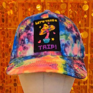 Op de afbeelding: Een tie-dye baseballcap met een levendige mix van kleuren, waaronder blauw, roze en geel. De cap heeft een zwart embleem met een paddenstoelafbeelding en de woorden "LET'S TAKE A TRIP!" in wit en roze.