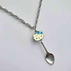 Op de afbeelding: Zilverkleurige ketting met een lepelvormige hanger met een Hello Kitty-ontwerp. De hanger heeft een lichtgeel gezicht, een blauwe strik en een kleine edelsteen. De ketting is in een gedraaide touwstijl.